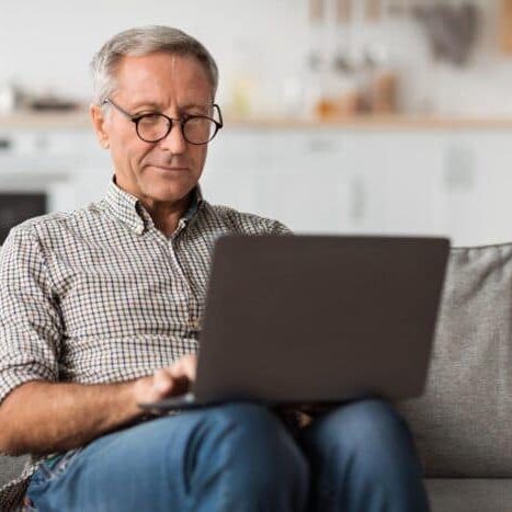 Senior Male Using Laptop Sitting On Couch At Home, Wearing Eyeglasses. Mature Freelancer Working Online And Browsing Internet On Computer In Living Room. Technology, Modern Retirement Lifestyle